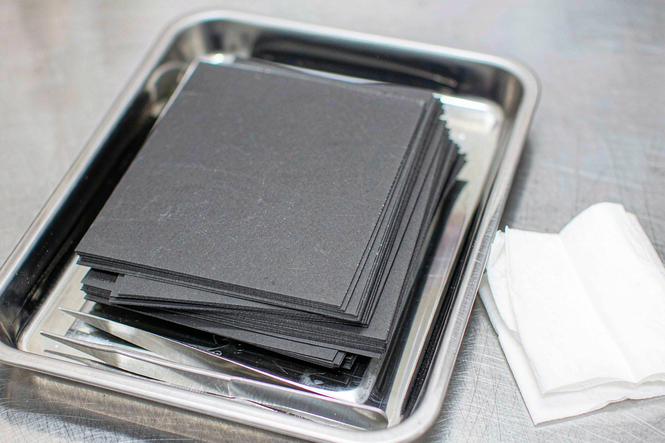 Semi-solid state battery moisture and contamination control: matte electrode sheets in a brushed stainless steel tray with tweezers and a lint-free wipe on a clean assembly bench